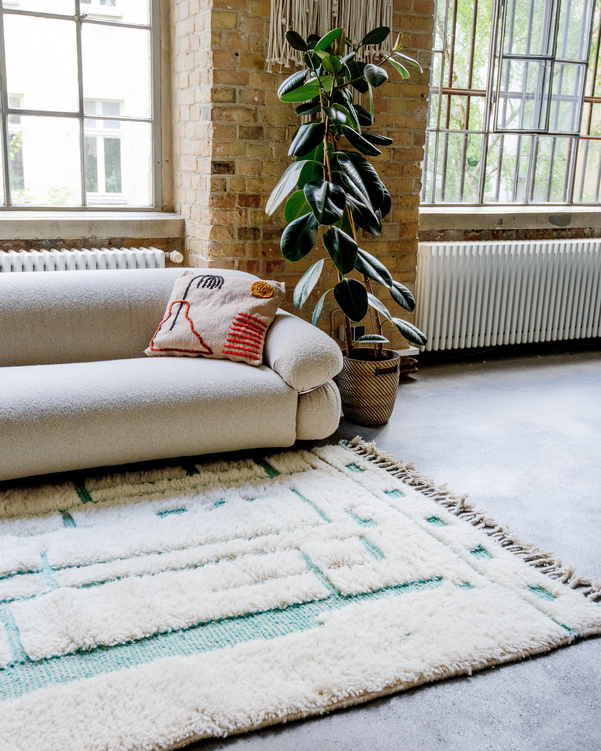 Cream rug with turquoise design lays on the floor in front of a cream couch and leafy plant.
